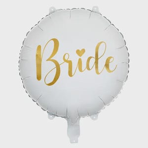 May include: A round white foil balloon featuring the word Bride written in a metallic gold cursive script font. A small gold heart is positioned above the letter i in the word. The balloon has a smooth surface with a crimped edge and a valve.