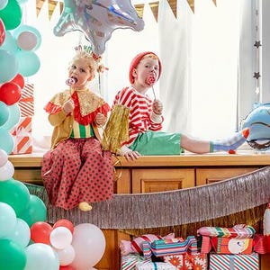 Puede incluir: Dos ni&ntilde;os con ropa festiva est&aacute;n sentados en un borde de madera, rodeados de globos coloridos y regalos envueltos. Un ni&ntilde;o lleva una capa de lentejuelas doradas y pantalones de lunares rojos, el otro un gorro rojo y una camiseta a rayas.