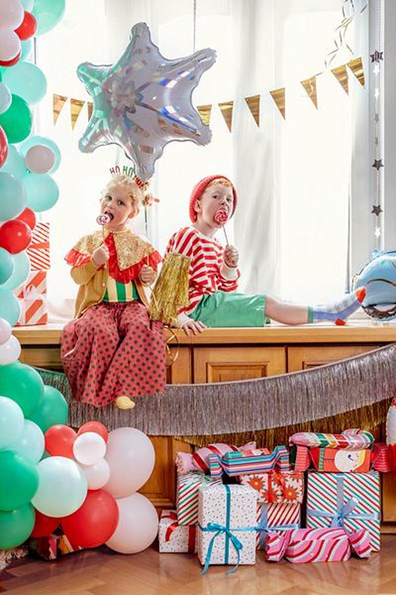 Puede incluir: Dos ni&ntilde;os con ropa festiva est&aacute;n sentados en un borde de madera, rodeados de globos coloridos y regalos envueltos. Un ni&ntilde;o lleva una capa de lentejuelas doradas y pantalones de lunares rojos, el otro un gorro rojo y una camiseta a rayas.