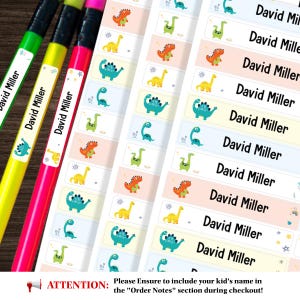 May include: A collection of rectangular adhesive labels featuring the name David Miller in black text alongside colourful illustrations of various dinosaurs. The labels are displayed on sheets and applied to three pencils in green, yellow, and pink.