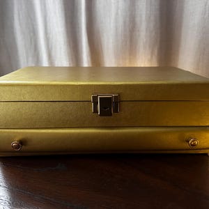May include: A gold-coloured jewellery box with a rectangular shape. The box has a textured surface and a metal clasp closure. Two small, round knobs are on the front. The box is closed and sits on a dark wooden surface.