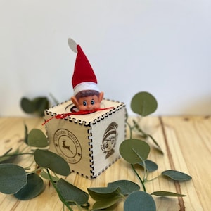 Peut inclure: Une boîte en bois avec un motif découpé au laser, avec un elfe qui regarde. L'elfe porte un chapeau rouge et blanc. La boîte porte un tampon circulaire avec un cerf et du texte. La verdure entoure la boîte.