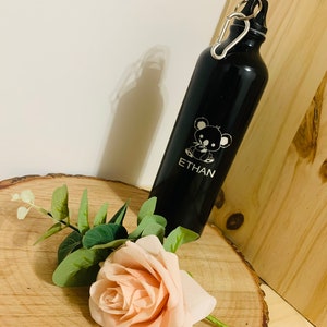 Puede incluir: Botella de agua reutilizable negra con un mosquetón plateado. La botella tiene un diseño de koala blanco y el nombre "ETHAN" grabado.