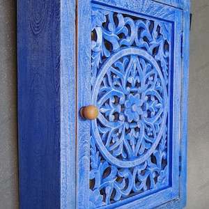 May include: A blue wooden cabinet with a carved floral design on the door. The cabinet has a single door with a wooden knob and is mounted on a wall.