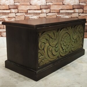 May include: A dark brown wooden storage chest with a carved green floral design on the front. The chest has a hinged lid and a decorative base.