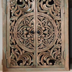 May include: A wooden cabinet with two doors featuring intricate carved floral designs. The cabinet is painted in a distressed green and brown finish.