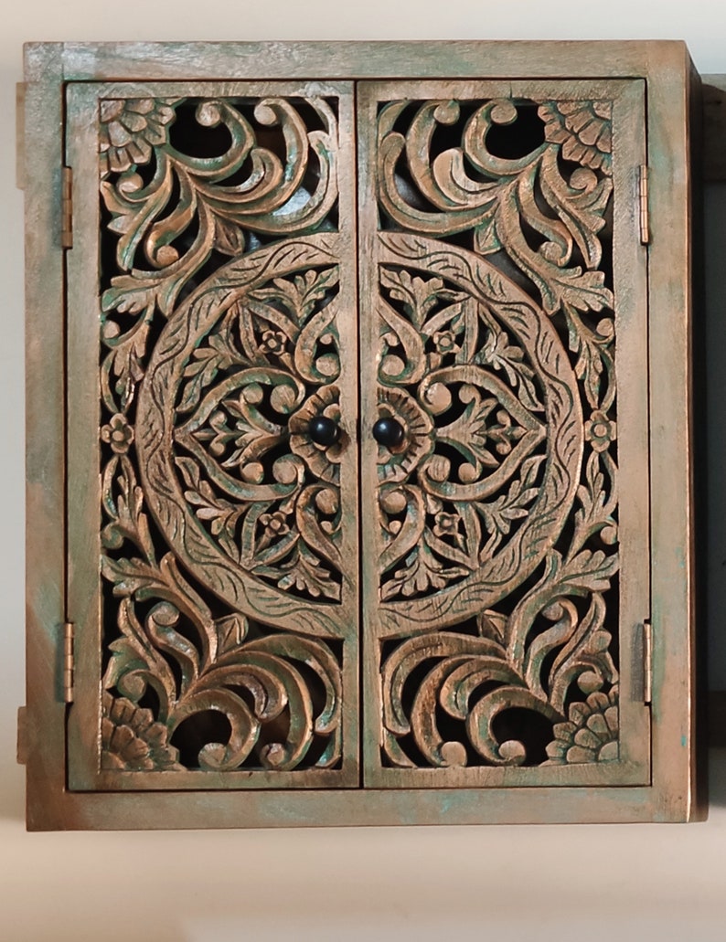 May include: A wooden cabinet with two doors featuring intricate carved floral designs. The cabinet is painted in a distressed green and brown finish.
