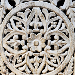 May include: White wooden wall decor with a carved circular design featuring intricate floral patterns. The design is symmetrical and features a central flower with multiple petals.
