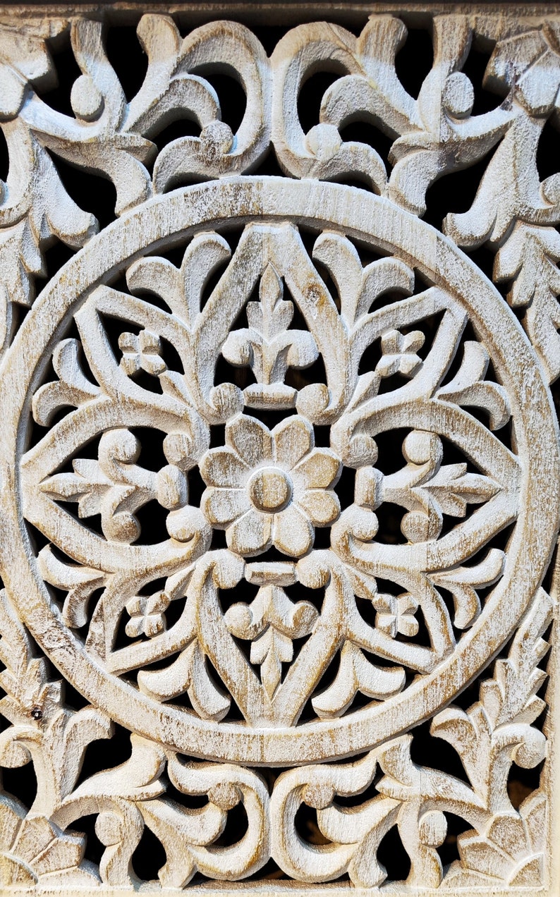 May include: White wooden wall decor with a carved circular design featuring intricate floral patterns. The design is symmetrical and features a central flower with multiple petals.