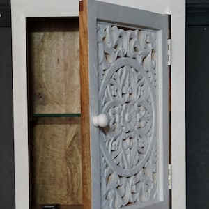May include: White wooden wall cabinet with a carved wooden door. The door is open and reveals a shelf inside. The cabinet has a simple knob.