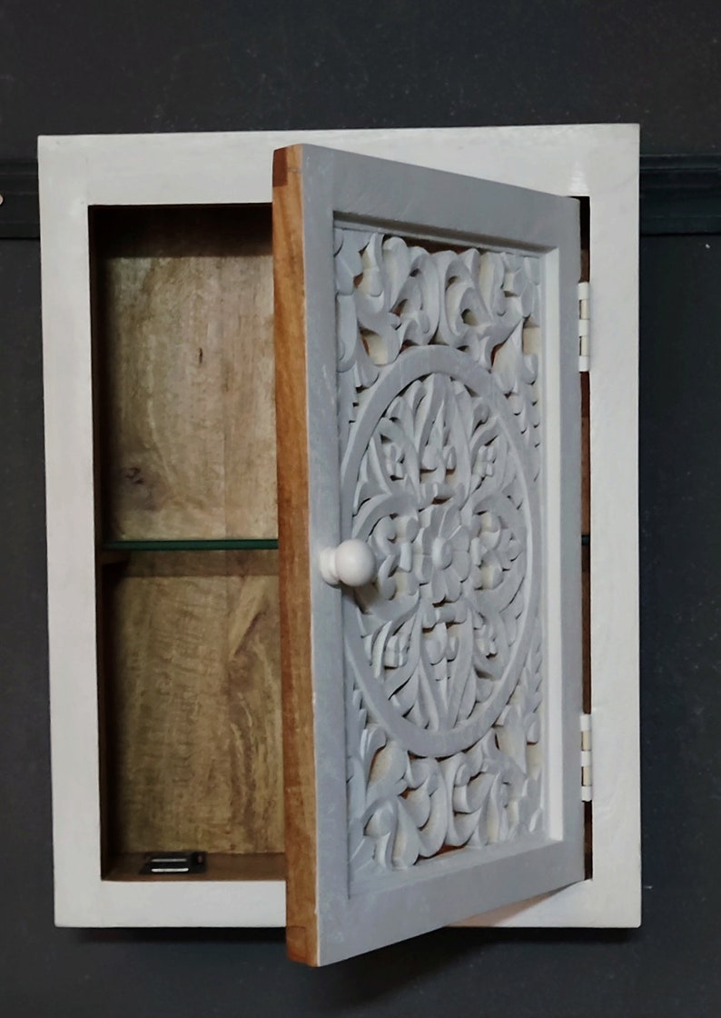 May include: White wooden wall cabinet with a carved wooden door. The door is open and reveals a shelf inside. The cabinet has a simple knob.