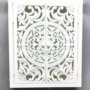 May include: White wooden cabinet with two doors featuring intricate carved floral designs. The doors have a circular pattern in the center and are closed.