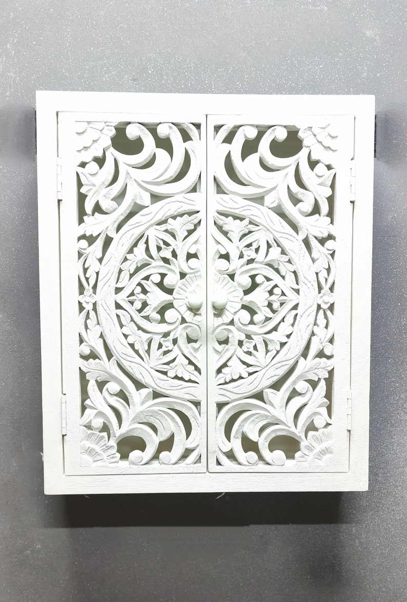 May include: White wooden cabinet with two doors featuring intricate carved floral designs. The doors have a circular pattern in the center and are closed.