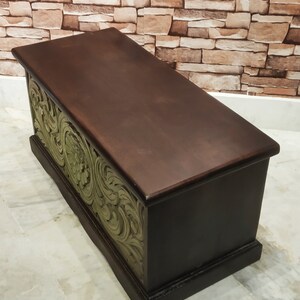May include: A dark brown wooden storage chest with intricate green floral carvings on the front. The chest has a hinged lid and is sitting on a light grey tiled floor.