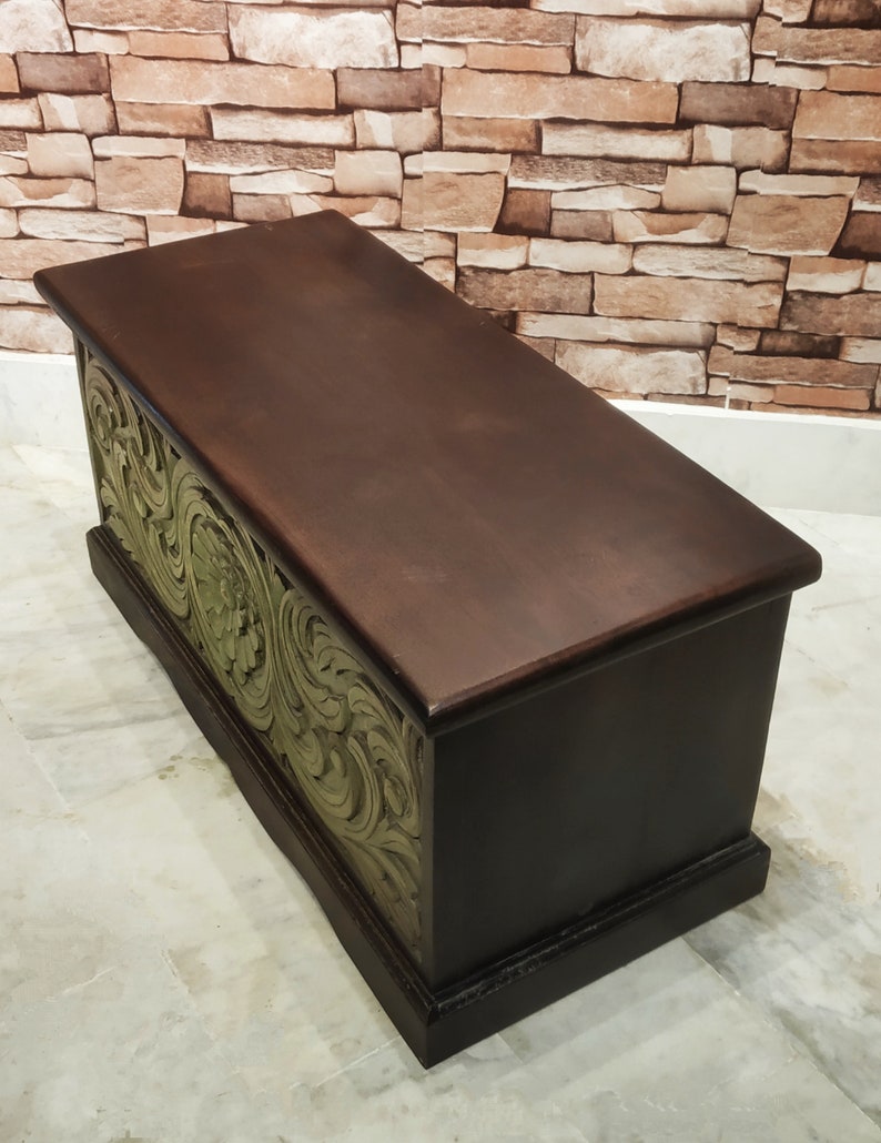 May include: A dark brown wooden storage chest with intricate green floral carvings on the front. The chest has a hinged lid and is sitting on a light grey tiled floor.