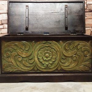 May include: A wooden storage chest with a carved floral design in green and brown. The lid is open, revealing the interior of the chest.