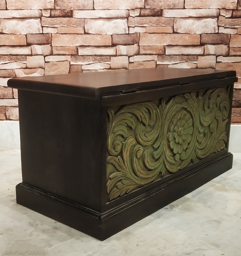 May include: A dark brown wooden storage chest with a carved green floral design on the front. The chest has a hinged lid and a decorative base.