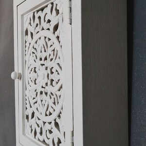 May include: White wooden cabinet with a carved, ornate door. The cabinet has a single knob and is mounted on a wall.