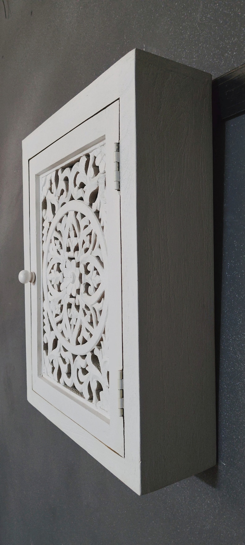 May include: White wooden cabinet with a carved, ornate door. The cabinet has a single knob and is mounted on a wall.