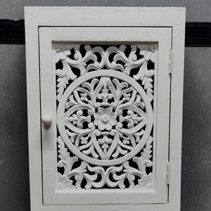 May include: White wooden cabinet with a carved floral design on the door. The cabinet has a single door and a small knob.