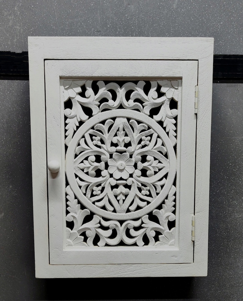 May include: White wooden cabinet with a carved floral design on the door. The cabinet has a single door and a small knob.