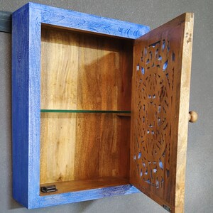 May include: A small wooden cabinet with a blue painted frame and a carved wooden door. The cabinet has one shelf and a glass front. The door has a knob and is open.