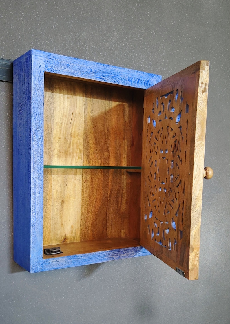May include: A small wooden cabinet with a blue painted frame and a carved wooden door. The cabinet has one shelf and a glass front. The door has a knob and is open.