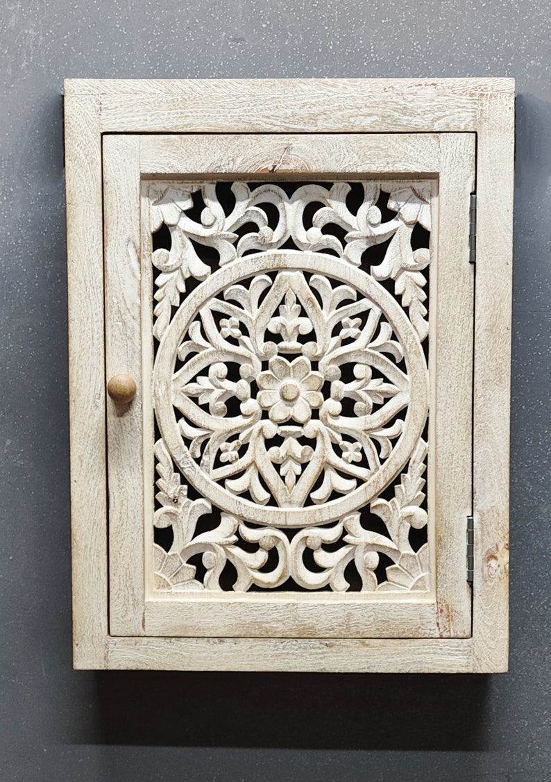 May include: White wooden cabinet with a carved floral design on the door. The cabinet has a single door with a knob and hinges.