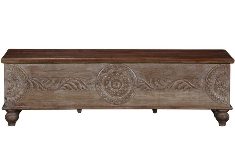 May include: A large, rectangular wooden storage chest with intricate carvings. The chest has a light brown finish and features a decorative floral design. It has four turned legs.