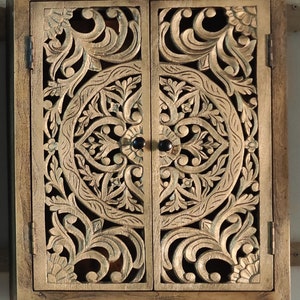 May include: A wooden cabinet with two doors featuring intricate carved floral designs. The doors are closed and have black metal knobs. The cabinet is painted in a light brown color.