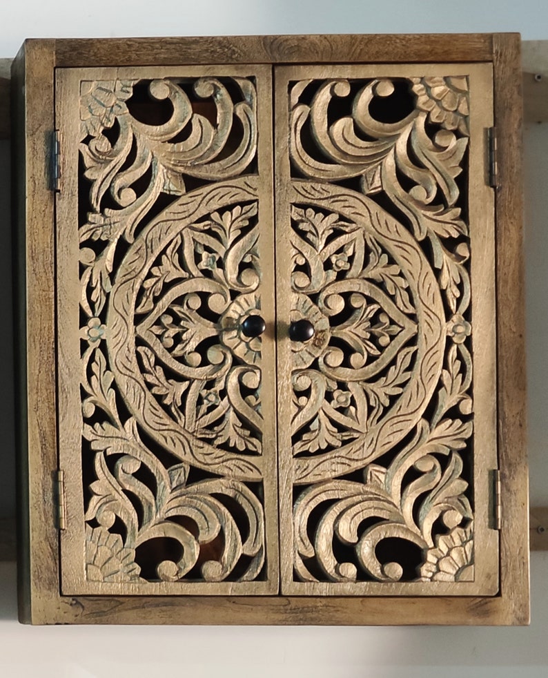 May include: A wooden cabinet with two doors featuring intricate carved floral designs. The doors are closed and have black metal knobs. The cabinet is painted in a light brown color.
