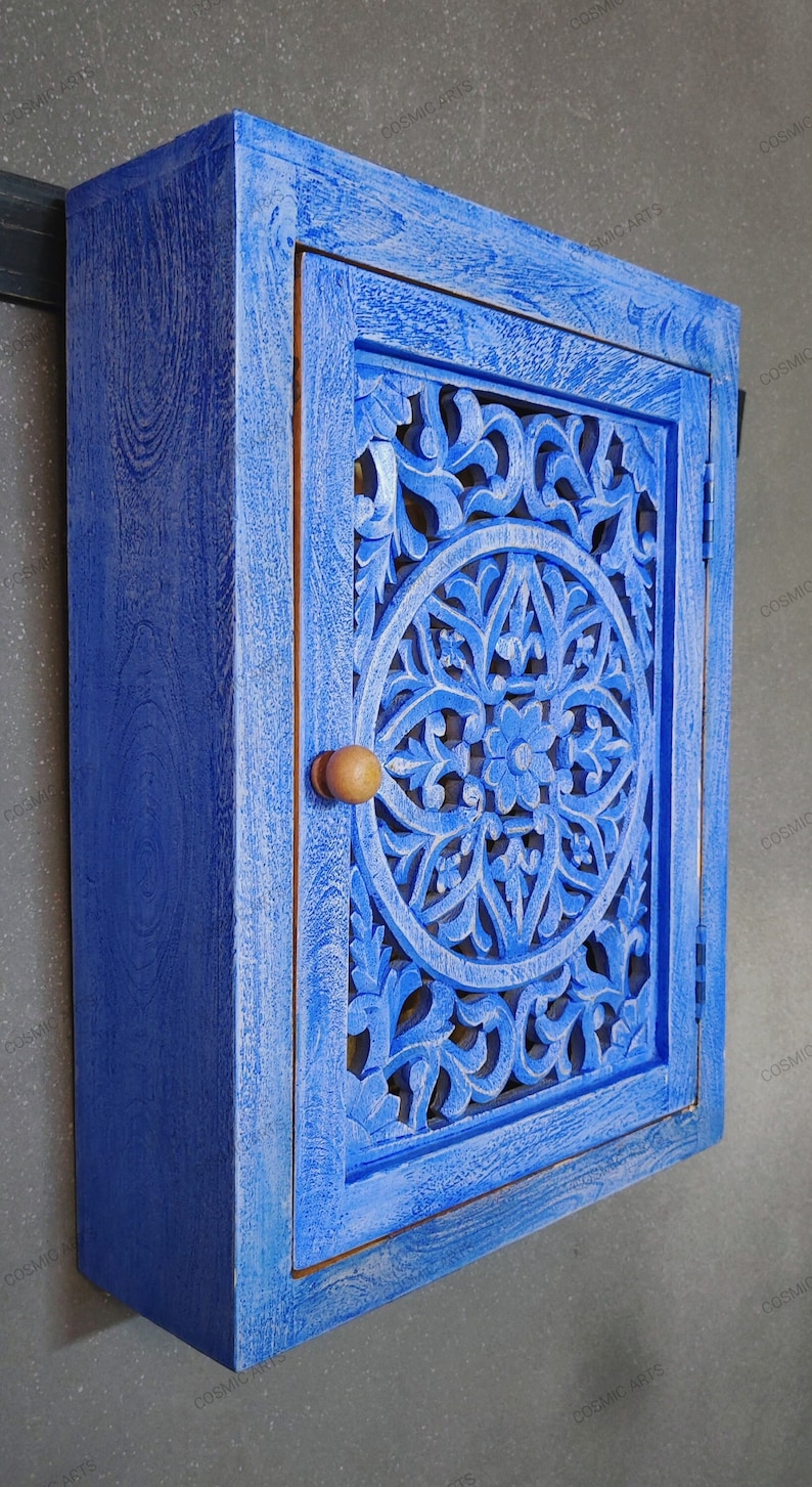 May include: A blue wooden cabinet with a carved floral design on the door. The cabinet has a single door with a wooden knob and is mounted on a wall.