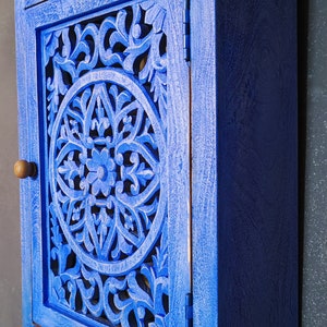 May include: A small blue wooden cabinet with a carved floral design on the door. The cabinet has a single door with a wooden knob and hinges.