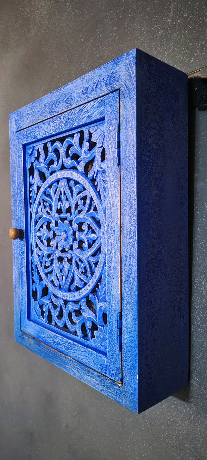 May include: A small blue wooden cabinet with a carved floral design on the door. The cabinet has a single door with a wooden knob and hinges.