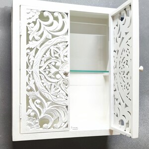 May include: White wooden wall cabinet with a carved floral design on the doors. The cabinet has one shelf and a glass door.