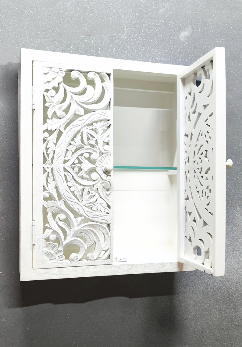 May include: White wooden wall cabinet with a carved floral design on the doors. The cabinet has one shelf and a glass door.
