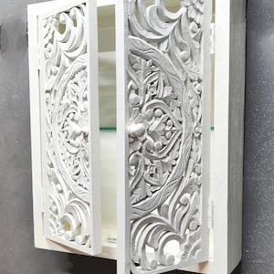May include: White wooden cabinet with intricate carved floral designs on the doors. The cabinet has a single shelf and a small knob on the door.