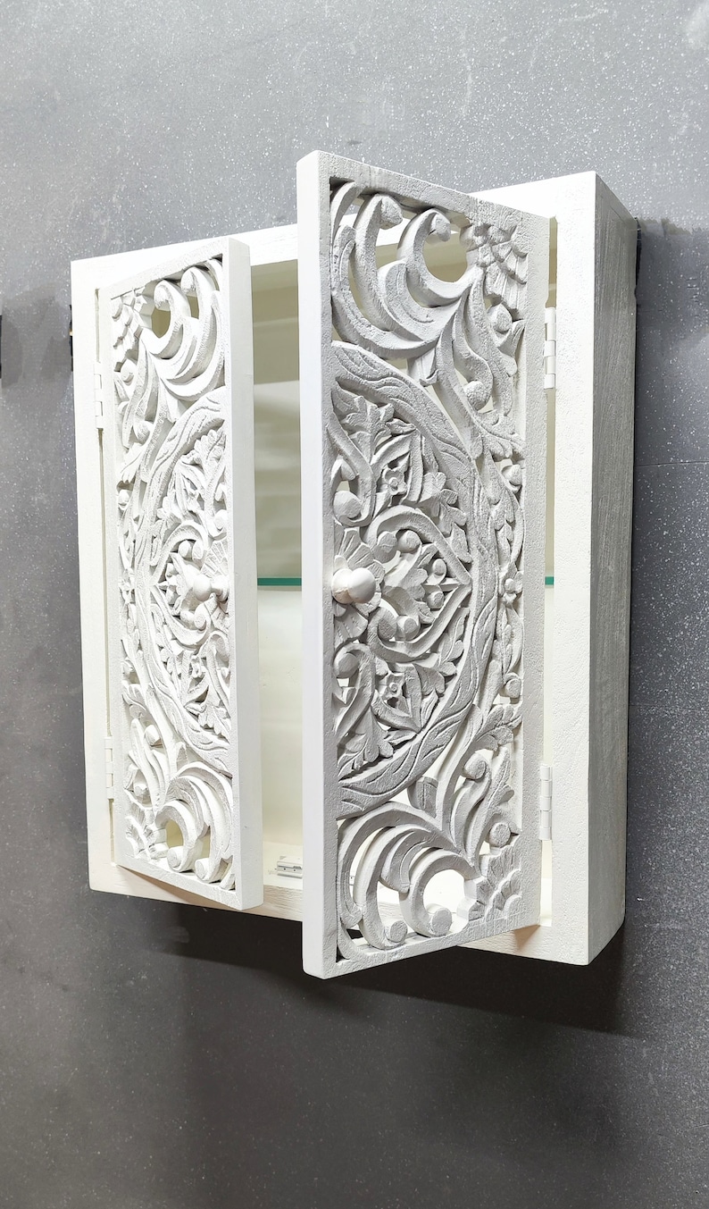 May include: White wooden cabinet with intricate carved floral designs on the doors. The cabinet has a single shelf and a small knob on the door.