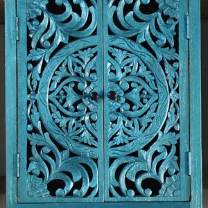 May include: A teal blue wooden cabinet with two doors. The doors are carved with intricate floral patterns and have a distressed finish.
