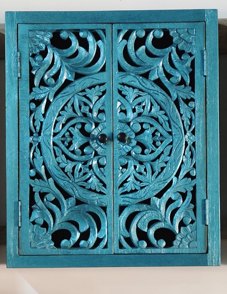 May include: A teal blue wooden cabinet with two doors. The doors are carved with intricate floral patterns and have a distressed finish.
