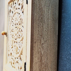 May include: A white wooden cabinet with a carved floral design on the door. The cabinet has a light brown finish and a simple knob.