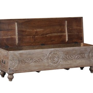 May include: A carved wooden storage bench with a light brown finish. The bench has a hinged lid and four turned legs. The lid is open, revealing the interior of the bench.