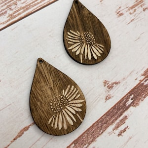 Puede incluir: Dos pendientes de madera en forma de lágrima con un diseño floral cortado con láser. Los pendientes son de color marrón oscuro con un diseño floral marrón claro.