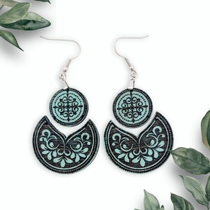 May include: A pair of wooden earrings with a teal and black design. The earrings are made of two pieces, a round piece with a floral design and a crescent-shaped piece with a more intricate floral design.