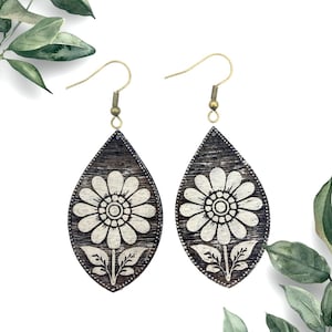May include: A pair of dangle earrings with a teardrop-shaped wooden base. The earrings are painted black with a white floral design. The earrings have a gold-tone hook.