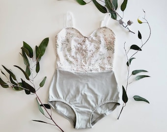 May include: A light gray one-piece swimsuit with a floral patterned bodice and thin straps. The swimsuit is surrounded by green eucalyptus branches on a white background.