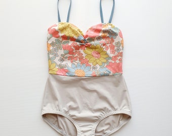 May include: A one-piece swimsuit with a floral top and a solid bottom. The top features a colorful floral pattern with yellow, pink, and blue flowers. The bottom is a solid light beige color. The straps are a light blue color.
