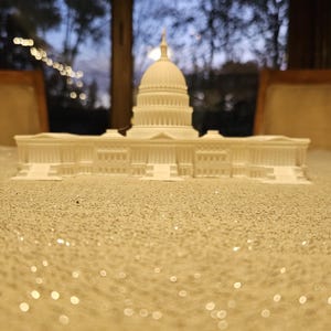 May include: A white miniature replica of the U.S. Capitol Building, featuring a domed roof and detailed architectural elements. The model is set on a glittery surface, with a blurred background of a window and trees.