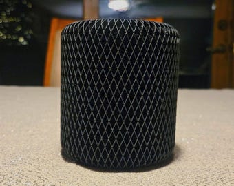 May include: A cylindrical black object with a diamond-patterned mesh design. The object is standing upright on a light-colored, textured surface. The background is blurred, suggesting an indoor setting.