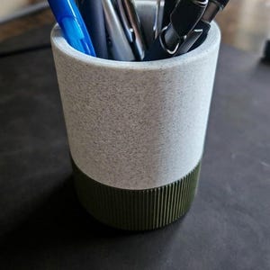May include: A cylindrical desk organizer with a speckled gray upper section and a ribbed, olive green base. The organizer holds various pens and pencils, including blue and black pens. The text "MODERN DESK ORGANIZER" is at the bottom.